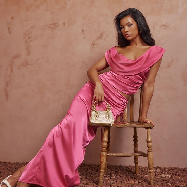 A woman sat on a stool, wearing a pink dress clutching a bag in her right hand